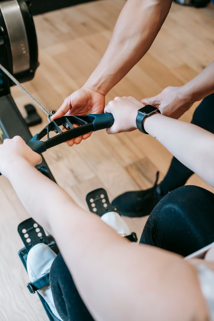 Home High angle of crop anonymous trainer assisting woman exercising on rowing machine in sports club